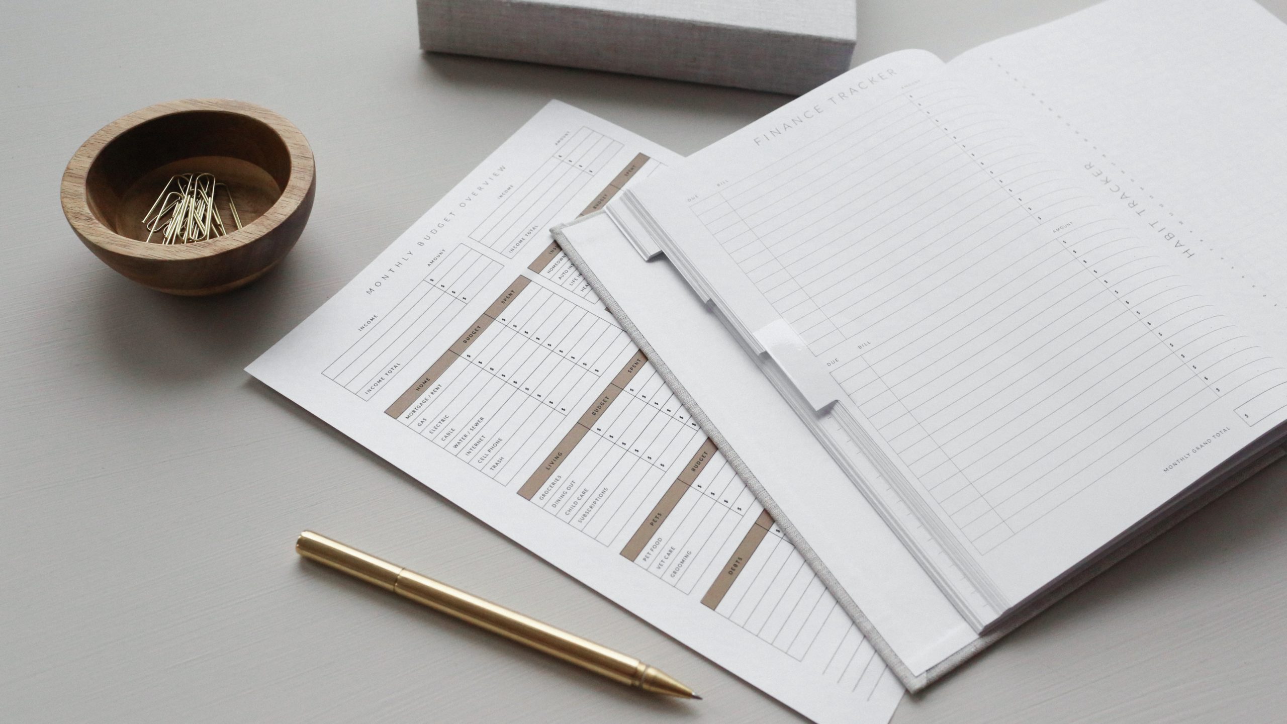Um livro de finanças e um diário estão abertos em uma mesa ao lado de uma caneta e uma tigela de clipes de papel.
