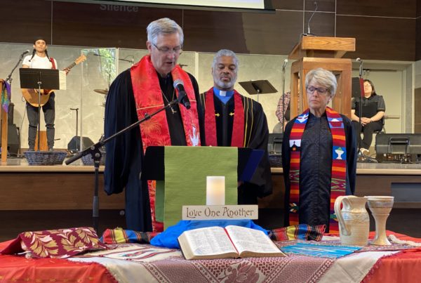 Phil Assink, Dwayne Jackson y Judy Nelson en el culto de clausura detrás de una mesa con la Cena del Señor, la Biblia y un cartel de "amaos los unos a los otros".